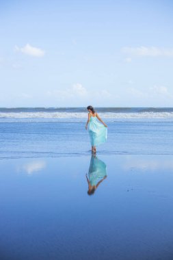 Genç bir kadın boş sahilde yalınayak yürüyor. Tam vücut portresi. İnce beyaz kadın, uzun elbise giyiyor. Arkadan bak. Su yansıması. Mavi gökyüzü. Asya 'da tatil. Seyahat konsepti. Bali, Endonezya