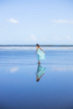 Genç bir kadın boş sahilde yalınayak yürüyor. Tam vücut portresi. İnce beyaz kadın, uzun elbise giyiyor. Arkadan bak. Su yansıması. Mavi gökyüzü. Asya 'da tatil. Seyahat konsepti. Bali, Endonezya