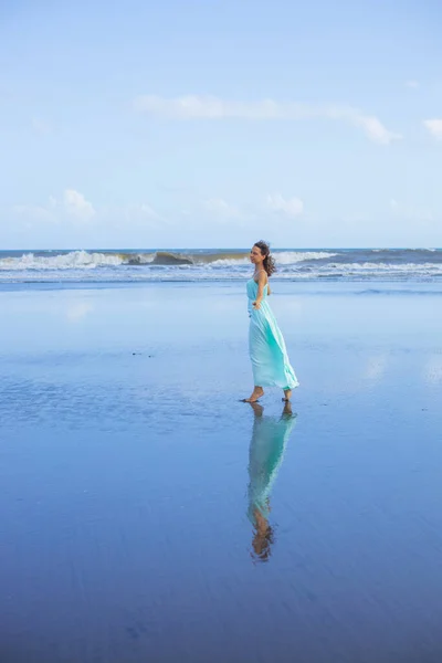 若い女性が空のビーチで裸足で歩く 全身肖像画 スリム白人女性は長いドレスを着ている 水の反射 夏の太陽の光 青い空だ アジアでの休暇 旅行のコンセプト バリ島インドネシア — ストック写真