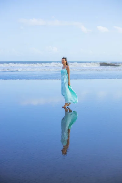 幸せな笑顔の女性が空のビーチで裸足で歩く 全身肖像画 スリム白人女性は長いドレスを着ている 水の反射 青い空だ アジアでの夏休み 旅行のコンセプト インドネシアのバリ — ストック写真