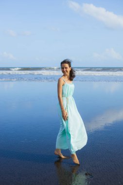 Boş sahilde çıplak ayakla yürüyen mutlu bir kadın. Tam vücut portresi. İnce beyaz kadın, uzun elbise giyiyor. Su yansıması. Mavi gökyüzü. Asya 'da yaz tatili. Seyahat konsepti. Bali, Endonezya