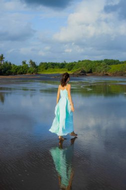 Sıska kadın boş sahilde yalınayak yürüyor. Tam vücut portresi. Beyaz kadın uzun elbise giyiyor. Arkadan bak. Su yansıması. Mavi bulutlu gökyüzü. Asya 'da tatil. Seyahat konsepti. Bali, Endonezya