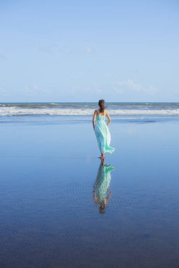 Genç bir kadın boş sahilde yalınayak yürüyor. Tam vücut portresi. İnce beyaz kadın, uzun elbise giyiyor. Arkadan bak. Su yansıması. Mavi gökyüzü. Asya 'da tatil. Seyahat konsepti. Bali, Endonezya