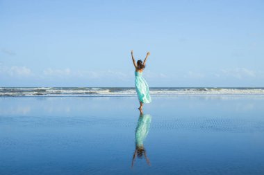Sıska kadın boş sahilde yalınayak yürüyor. Eller yukarı! Beyaz kadın uzun elbise giyiyor. Arkadan bak. Su yansıması. Mavi gökyüzü. Asya 'da tatil. Özgürlük kavramı. Bali, Endonezya