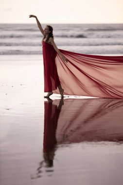 Gün batımında kumsalda dans eden zayıf bir kadın. Su yansıması. Uzun kırmızı elbise giyen güzel beyaz kadın. Mükemmel vücut şekli. Romantik bir kavram. Asya 'ya git. Bali, Endonezya