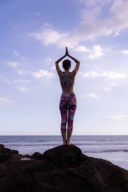 Kayanın üzerinde durmuş yoga yapan bir kadın. Genç bir kadın plajda namaste Mudra ile kollarını kaldırıyor. Mavi gökyüzü arka planı. Bali 'de yoga inzivası. Arkadan bak. Dikey yerleşim. Uzayı kopyala Endonezya