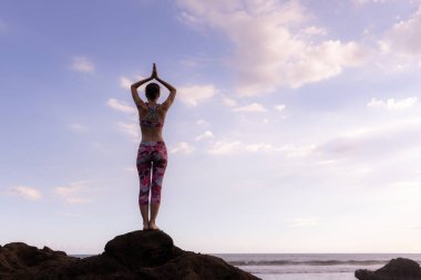 Kayanın üzerinde durmuş yoga yapan bir kadın. Genç bir kadın plajda namaste Mudra ile kollarını kaldırıyor. Mavi gökyüzü arka planı. Yoga inzivası. Arkadan bak. Yatay yerleşim planı. Uzayı kopyala Bali, Endonezya