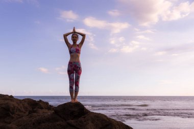 Sıska kadın kayanın üzerinde duruyor, sahilde yoga yapıyor. Namaste Mudra 'da eller havaya. Mavi gökyüzü arka planı. Yoga inzivası. Konsantrasyon ve denge. Yatay yerleşim planı. Uzayı kopyala Bali