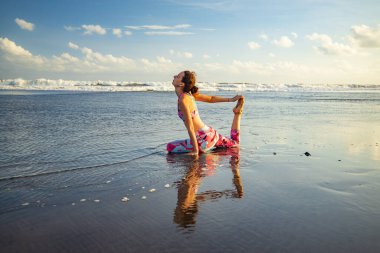 Plaj yogası Eka Pada Rajakapotasana, Tek Ayaklı Kral Güvercin Pozu pratiği yapan kadın. Göğüs açıcı nefes almakta güçlük çekiyor. Kişisel bakım konsepti. Sağlıklı bir hayat. Yoga inzivası. Su yansıması. Bali, Endonezya