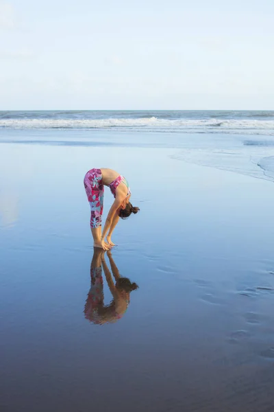 ビーチでの太陽の敬礼練習 ウッタナサナに立つスリムな女性 前かがみに立つ 健康的なライフスタイル 柔軟な背骨だ 水の反射 ヨガの隠れ家だ コピースペース インドネシアのバリ — ストック写真
