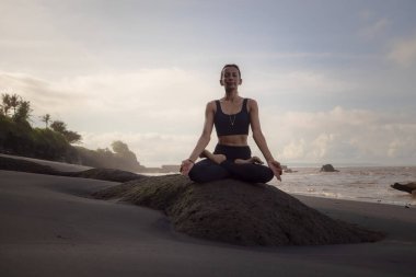 Sahilde meditasyon yogası. Lotus pozunda kayanın üzerinde oturan Asyalı kadın. Padmasana. Eller Gyan Mudra 'da. Yoga inzivası. Kapalı gözler. Sağlıklı bir kavram. Uzayı kopyala Mengening Plajı, Bali, Endonezya