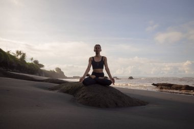 Sahilde sabah meditasyonu ve yoga. Lotus pozunda kayanın üzerinde oturan Asyalı kadın. Padmasana. Eller Gyan Mudra 'da. Yoga inzivası. Sağlıklı bir kavram. Uzayı kopyala Mengening Plajı, Bali, Endonezya