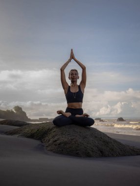 Sahilde meditasyon yogası. Lotus pozunda kayanın üzerinde oturan Asyalı kadın. Padmasana. Namaste Mudra 'da eller havaya. Yoga inzivası. Sağlıklı bir kavram. Uzayı kopyala Mengening Plajı, Bali, Endonezya
