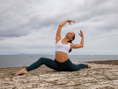Genç bir kadın bacak açma alıştırması yapıyor. Esnek vücut. Açık hava egzersizi. Fitness, spor, jimnastik, sağlık konsepti. Sağlıklı bir yaşam tarzı. Bulutlu bir arka plan. Uzayı kopyala Bali Endonezya