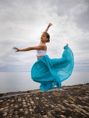 Bulutlu gökyüzünden atlayan güzel genç bir kadın. Beyaz kadın, mavi uzun etek giyiyor. Bale dansı konsepti. Açık hava aktivitesi. Uzayı kopyala Gökyüzü arka planı. Bali, Endonezya