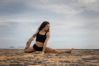 Sportif ve atletik genç bir bayan bacağını ayırıyor. Kumsalda esneme egzersizi. Spor açık hava konsepti. Sağlıklı bir yaşam tarzı. Beyaz kadın siyah spor giysisi giyiyor. Uzayı kopyala Bali, Endonezya