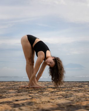 Beyazlı sporcu bir kadın plajda soyunuyor ve ısınıyor. İleriye doğru eğil. Esnek omurga. Sağlıklı aktif yaşam tarzı. Açık hava fitness. Sağlık konsepti. Uzayı kopyala Bali, Endonezya