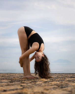 Beyazlı sporcu bir kadın plajda soyunuyor ve ısınıyor. İleriye doğru eğil. Esnek omurga. Sağlıklı aktif yaşam tarzı. Açık hava fitness. Sağlık konsepti. Uzayı kopyala Bali, Endonezya