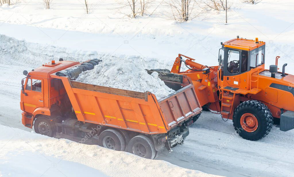 Big orange tractor cleans up snow from the road and loads it into the truck. Cleaning and cleaning of roads in the city from snow in winter