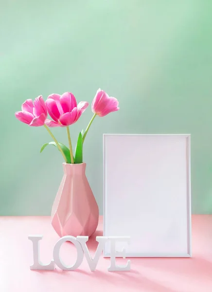 Рожеві тюльпани у вазі, порожня біла рамка, білі літери LOVE на пастельному рожево-зеленому тлі. Знущання . — стокове фото