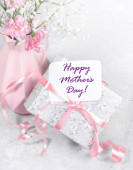 Zabalená květinovým papírem, dárková krabička zdobená stuhou a na ní nápis ke dni matek a růžová váza růžových karafiátů a gypsophily na světle šedém pozadí zblízka. Svislá orientace.