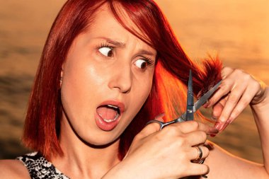 Şok olmuş kızıl saçlı bir kız dışarıda saçını makasla kesiyor. Kız kırık uçlarıyla şoka girdi..