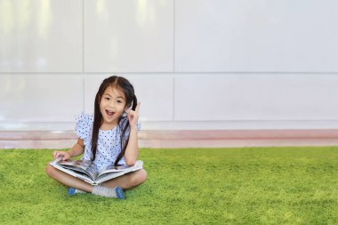 Asyalı özgüvenli küçük kızın portresi oturma odasında yeşil çimlerde oturan mutlu ve gülen yüzlü bir şeyi işaret ederek kitap ya da dergi okurken