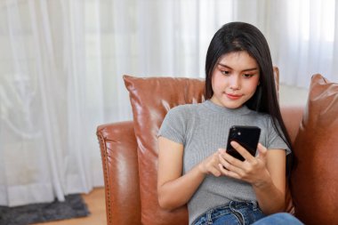 Genç zeki ve aktif Asyalı kadın oturma odasında koltukta oturuyor, mutlu yüzlü akıllı telefon kullanıyor.