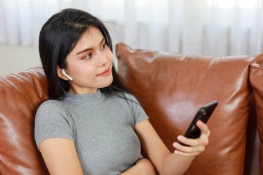 Genç zeki ve aktif Asyalı kadın oturma odasında koltukta oturuyor, mutlu yüzlü akıllı telefon kullanıyor.