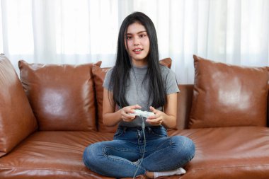 VDO oyun konsolu konsepti. Aktif Asyalı kadın kanepede oturuyor, elinde joystickle heyecan verici bir oyun oynuyor. Sevimli kız oyun kumandası konsolunda heyecanlı görünüyordu.