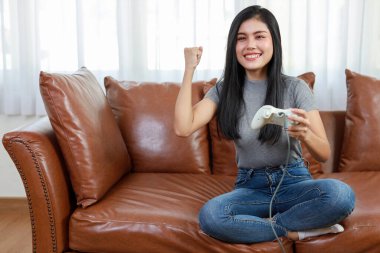 VDO oyun konsolu konsepti. Aktif Asyalı kadın kanepede oturuyor, elinde joystickle heyecan verici bir oyun oynuyor. Sevimli kız oyun kumandası konsolunda heyecanlı görünüyordu.