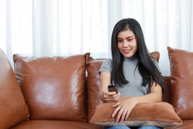 Televizyon ve mutluluk konsepti. Oturma odasındaki kanepede oturmuş elinde uzaktan kumandayla gülümseyen güzel bir Asyalı kadın. Yaşam tarzı kavramı.