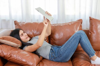 Genç zeki ve aktif Asyalı kadın oturma odasında kanepede uzanmış, mutlu yüzlü tablet kullanıyor, yaşam tarzı ve teknolojisi var.