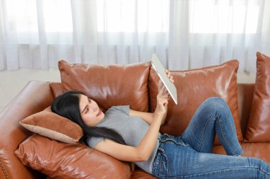 Genç zeki ve aktif Asyalı kadın oturma odasında kanepede uzanmış, mutlu yüzlü tablet kullanıyor, yaşam tarzı ve teknolojisi var.