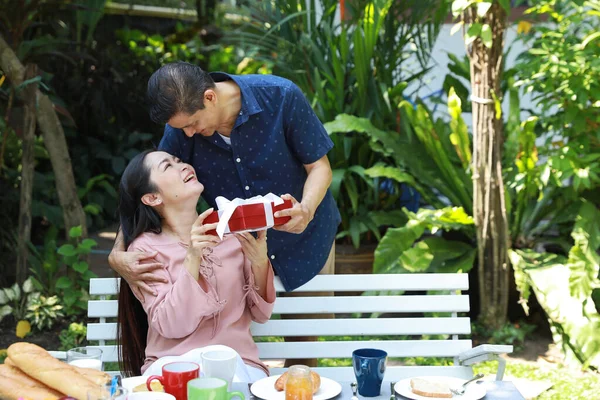 在结婚周年纪念日当天 亚洲的一对老夫妻坐在后院的一张早餐桌旁 亚洲年长的丈夫笑着送给亚洲年长的妻子惊喜的礼物 — 图库照片