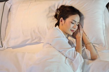 Beyaz uyku tulumu giyen Asyalı bir kadın yatakta yatarken ya da yatakta mutlu bir şekilde uyur ve güzel rüyalar görürken gülümserdi. Yaşam tarzı kavramı