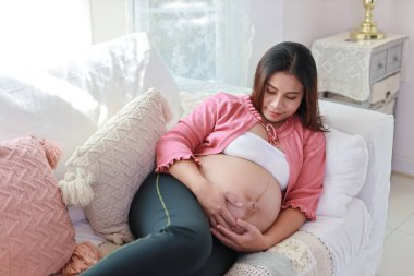 Mutlu gülümseyen Asyalı hamile kadın dinleniyor ve rahatlamaya çalışırken oturma odasında kanepede uzanıyor. Müstakbel anne hamilelik süresince doğum için hazırlanıyor ve bekliyor..