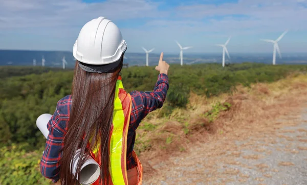 Windmill engineer young woman pointing and planing renewable energy technology or alternative ecology project for future. Electrician engineer looking and checking wind turbines site with blueprint