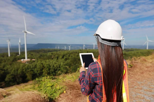 Windmill engineer young woman using tablet and planing renewable energy technology or alternative ecology project for future. Electrician engineer looking and checking wind turbines site with blue sky