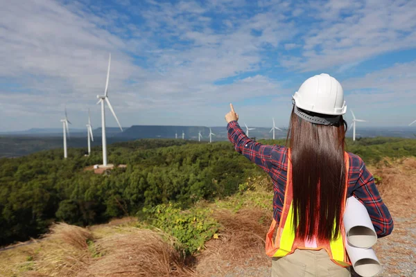 Windmill engineer young woman pointing and planing renewable energy technology or alternative ecology project for future. Electrician engineer looking and checking wind turbines site with blueprint