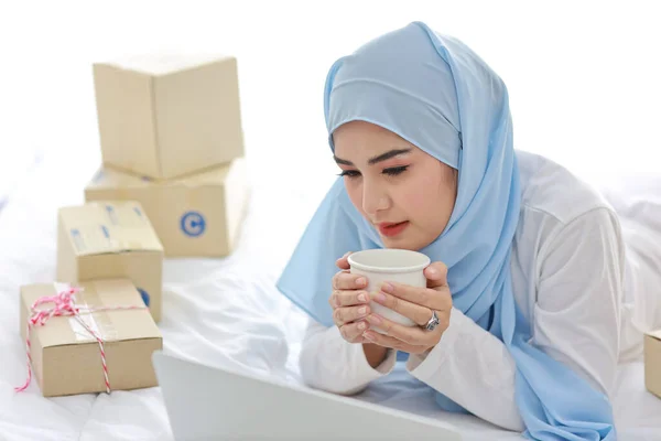 寝室の若い魅力的なアジアの女性は イスラム教徒のドレスを着て コンピュータとオンラインパッケージボックスの配信でベッドの上にコーヒーカップを保持 スタートアップ小企業携帯電話で働く中小企業フリーランスの女の子 — ストック写真
