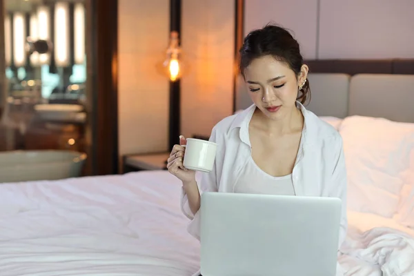 Happy asian woman in casual sitting on bed and holding coffee cup for breakfast while using computer in hotel, beauty businesswoman with bedroom background. Lifestyle technology concept