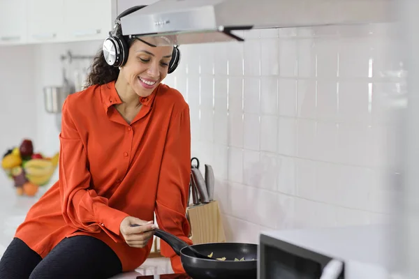 Happy latin woman cooking, smelling and tasting food in kitchen. Beautiful young female with headphone preparing delicious meal while listen music at home. Healthy and lifestyle concept