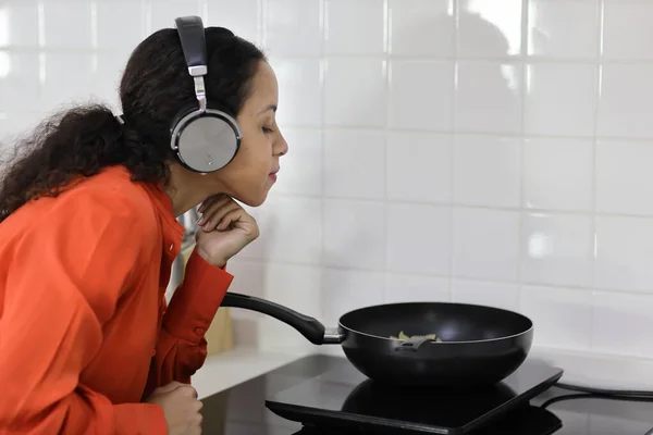 Happy latin woman cooking, smelling and tasting food while making meal in kitchen. Beautiful young female with headphone prepare delicious meal and listen music at home. Healthy and lifestyle concept
