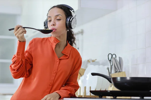 Happy latin woman cooking, smelling and tasting food with spoon in kitchen. Beautiful young female with headphone preparing delicious meal while listen music at home. Healthy and lifestyle concept
