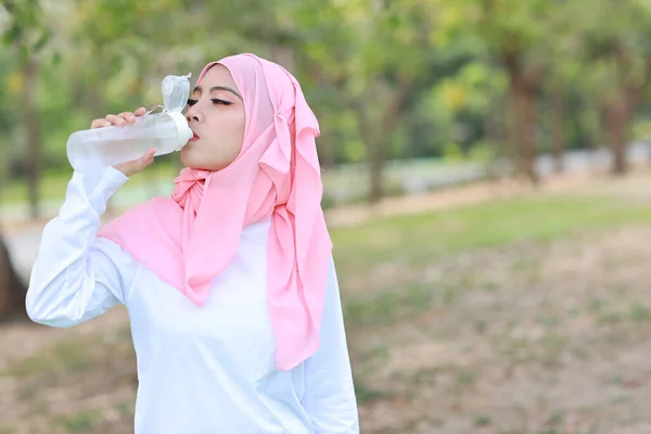 漂亮的健美运动员亚洲女人在运动后喝水 年轻可爱的女孩穿着穆斯林运动服 带着耳机 在户外运动后休息 体育概念 — 图库照片