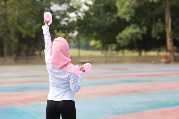 后视镜下 身穿穆斯林运动服的年轻亚洲女子站在室外举起哑铃进行晨练 活泼的女孩在有绿树背景的二头肌运动中锻炼 体育概念 — 图库照片