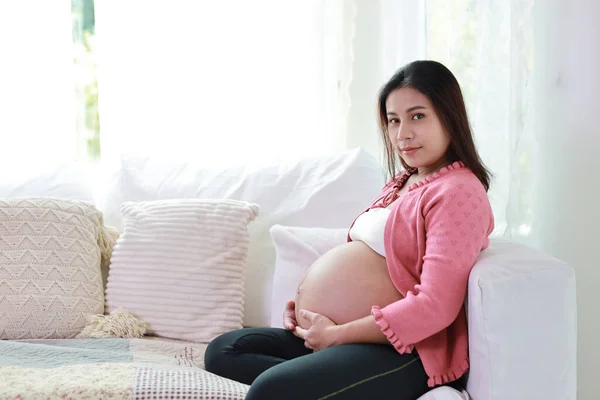 Happy smiling young asian pregnant woman resting and sitting on sofa in living room while touching and looking camera. Expectant mother preparing and waiting for baby birth during pregnancy.