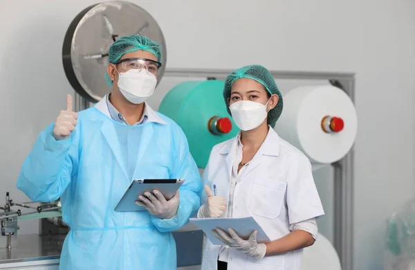 Workers in personal protective equipment or PPE inspecting holding tablet and clipboard while showing thumb up in face mask production line factory, manufacturing industry and factory concept.
