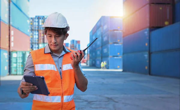 Foreman using walkie talkie radio and tablet control loading containers box. Engineer or worker with safety hat work at container cargo site and checking industrial container cargo freight ship